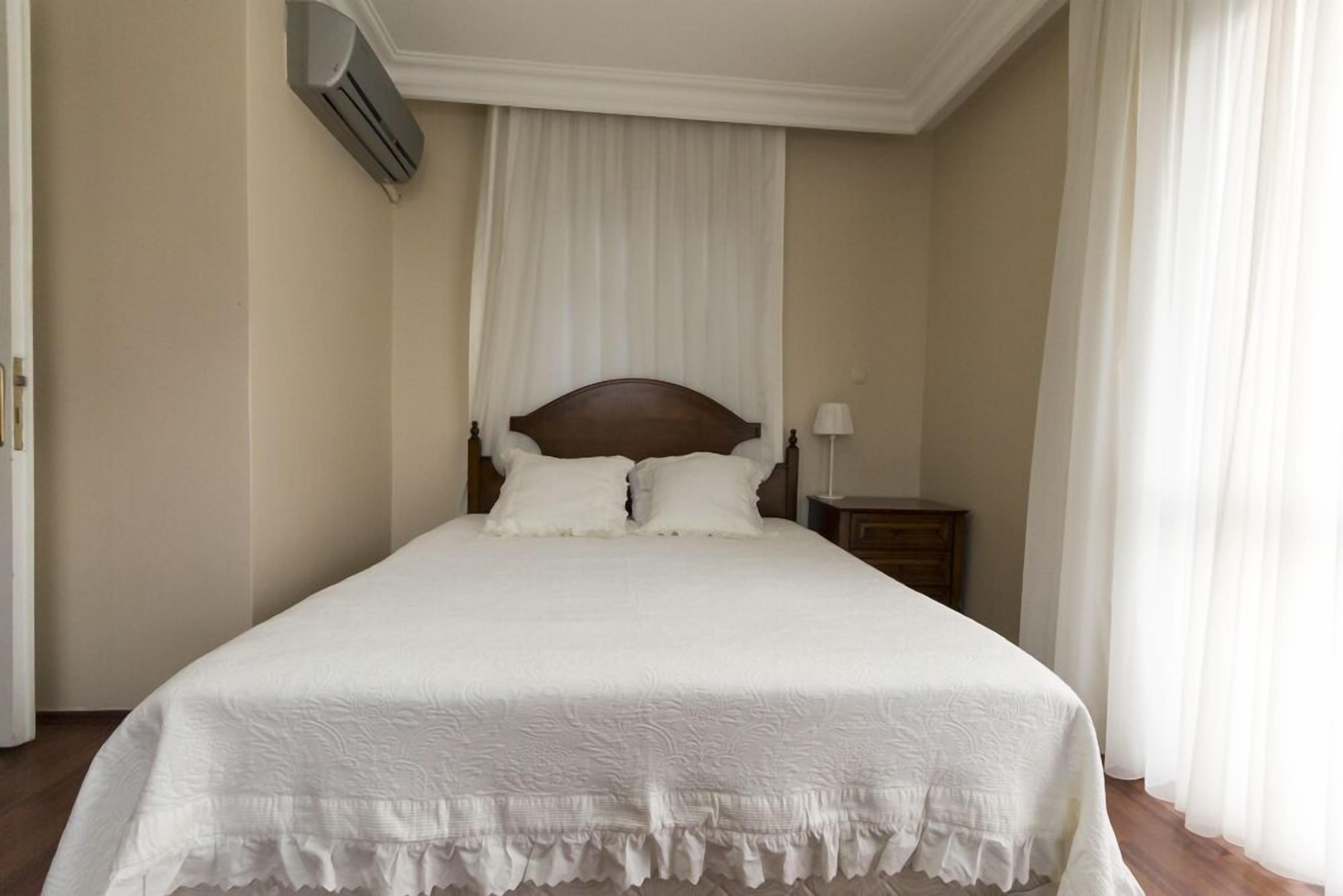 Una cama bien hecha con cabecera de madera, ropa de cama blanca y una mesita de noche con una lámpara.
