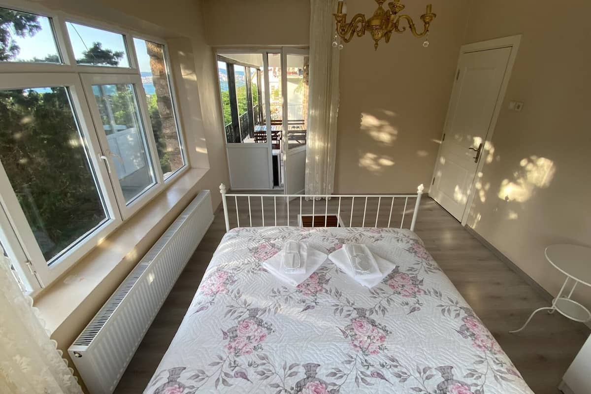 A bedroom with a large window, a radiator, a bed with a floral duvet, a chandelier, and a view of a balcony.