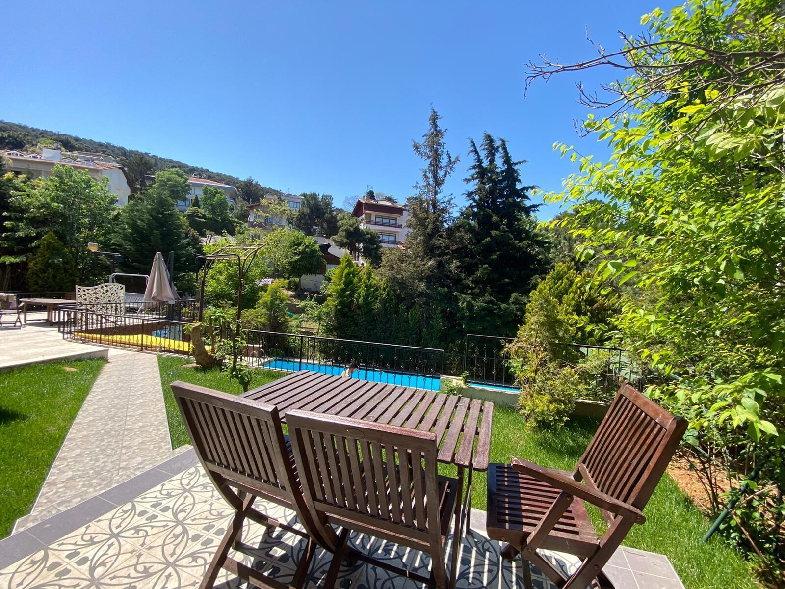 A patio with wooden tables and chairs, a swimming pool, and a garden with trees and a view of buildings in the background.