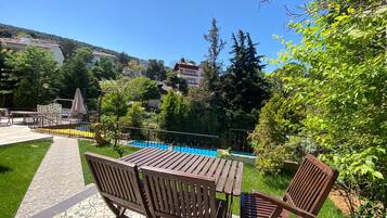 A patio with wooden tables and chairs, a swimming pool, and a garden with trees and a view of buildings in the background.