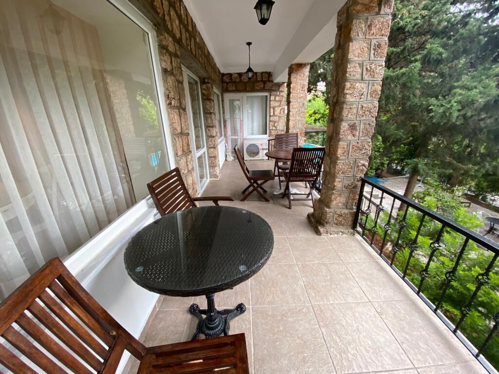 A balcony with a round table and wooden chairs, stone walls, and a view of greenery.