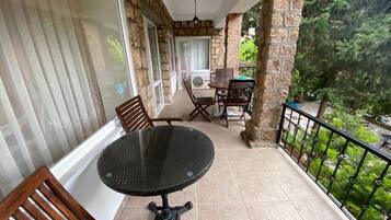 A balcony with a round table and wooden chairs, stone walls, and a view of greenery.