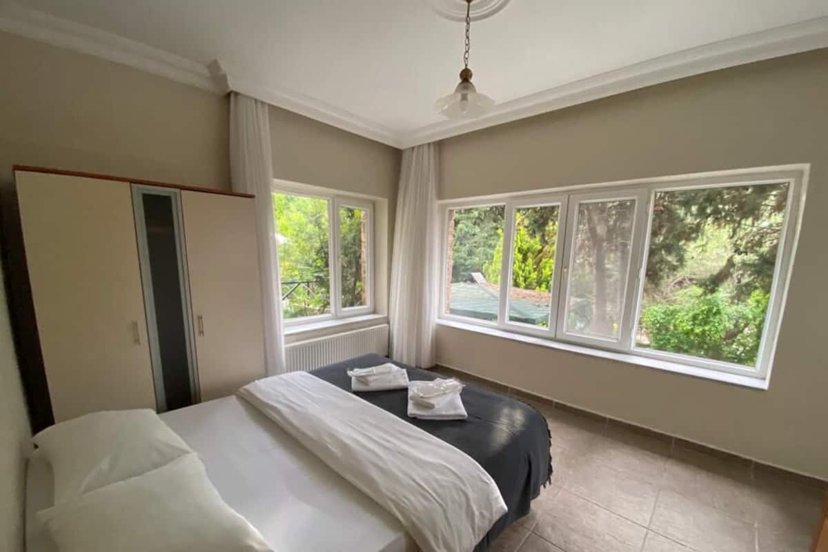 A bedroom with a bed, a wardrobe, and large windows with a view of greenery.