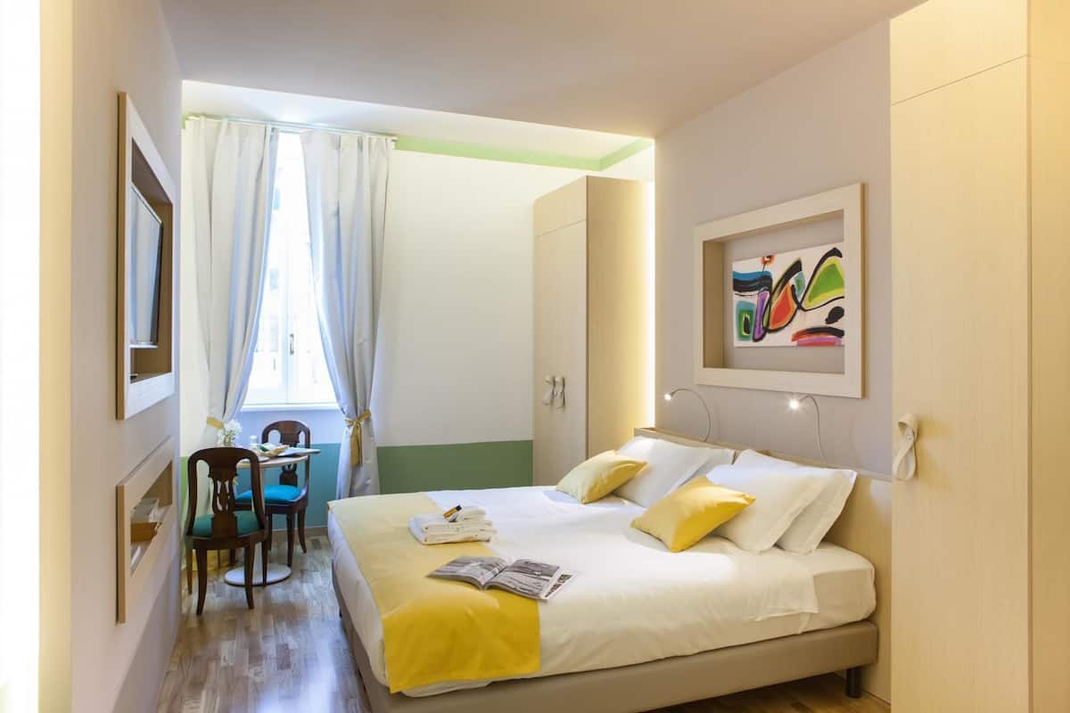 A hotel room with a bed, a desk, and a chair. There is a window with curtains, a painting on the wall, and a door leading to another room.