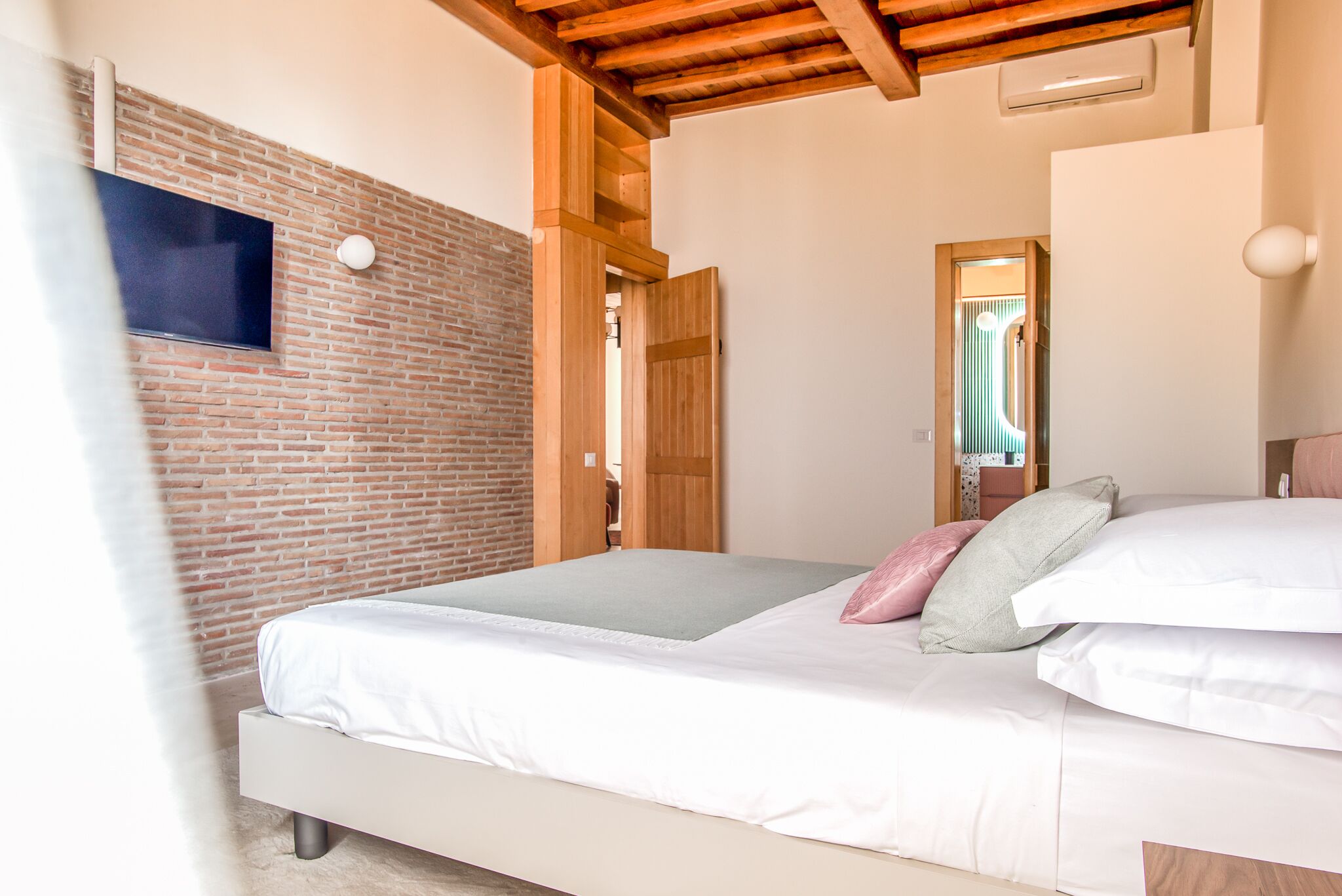 A bedroom with a brick wall, a wooden ceiling, a bed with white bedding, and a TV mounted on the wall.