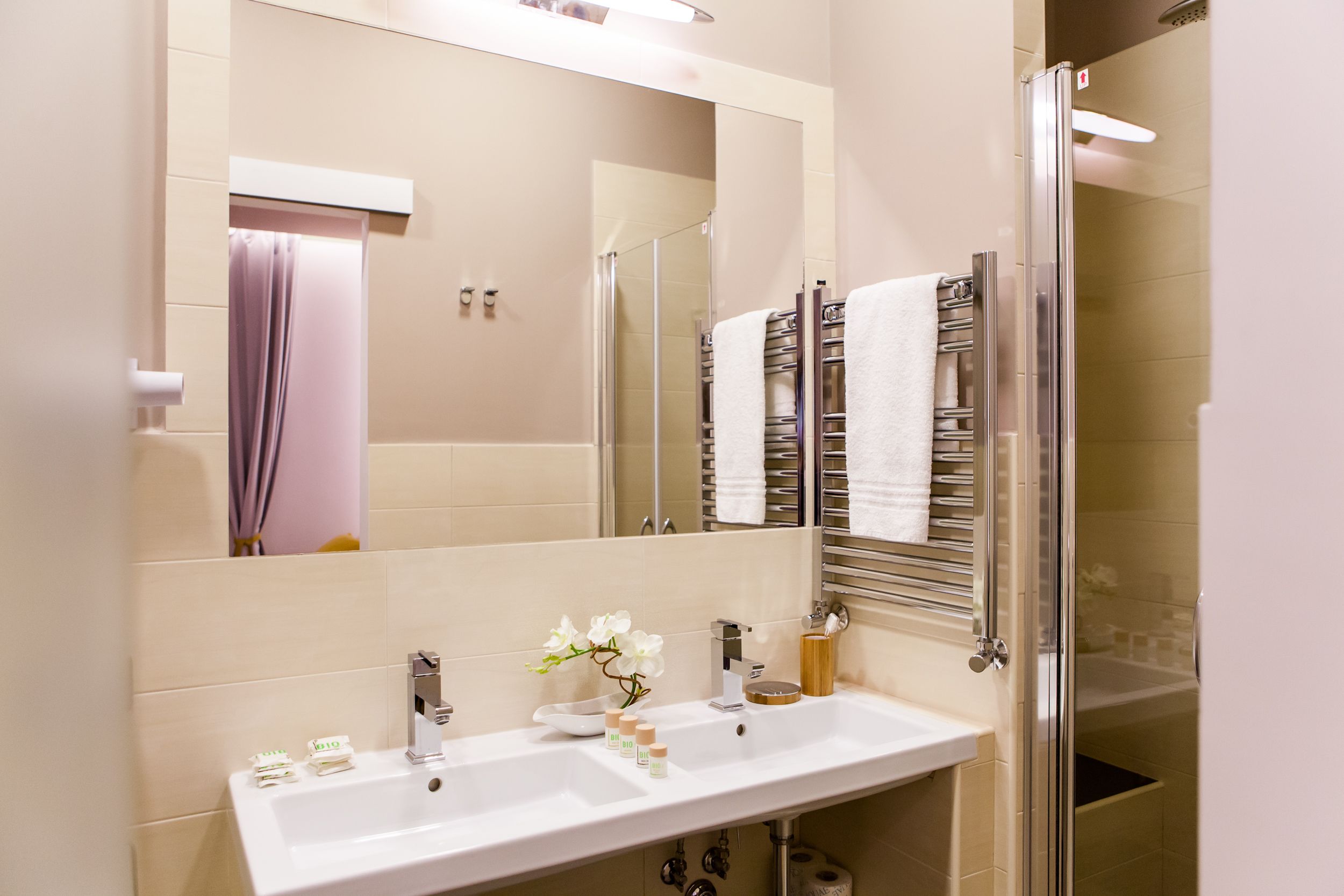 A modern bathroom with a large mirror, two sinks, a glass shower enclosure, and a towel rack with multiple towels.