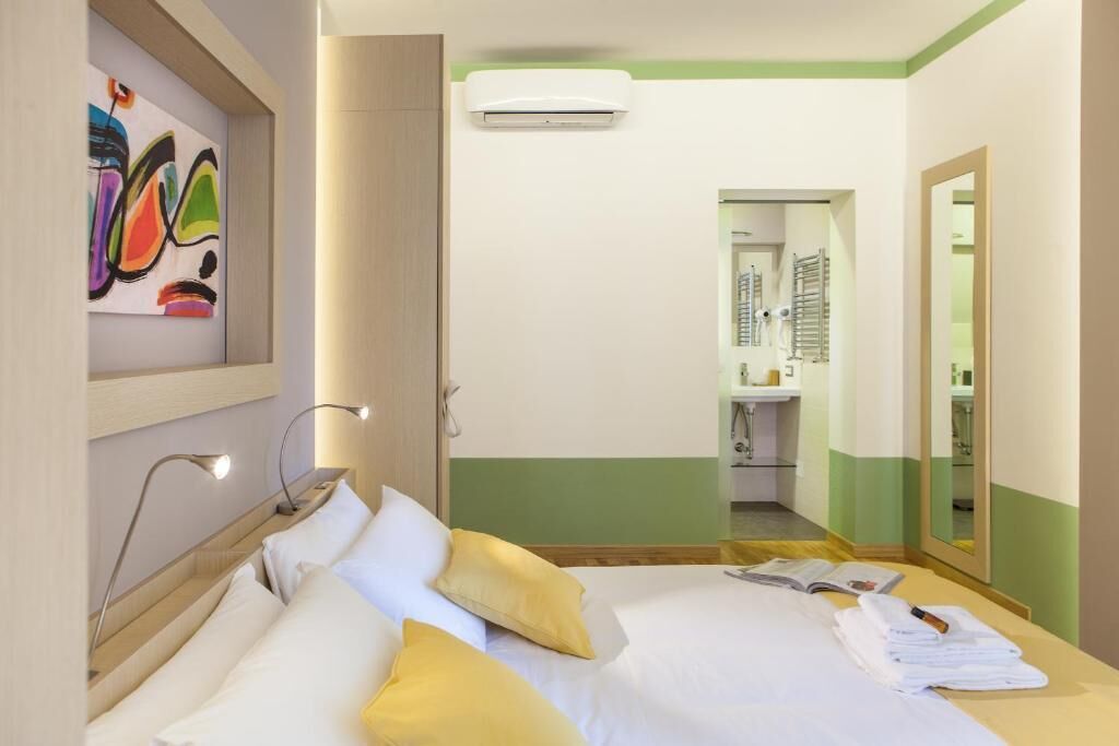 A hotel room with a bed, bedside lamps, a mirror, a wall-mounted air conditioner, and a bathroom visible through an open door.
