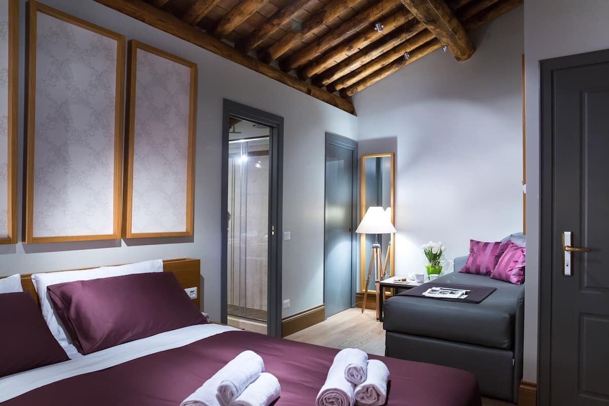 A hotel room with a bed, a sofa, a desk with a lamp, and a door.