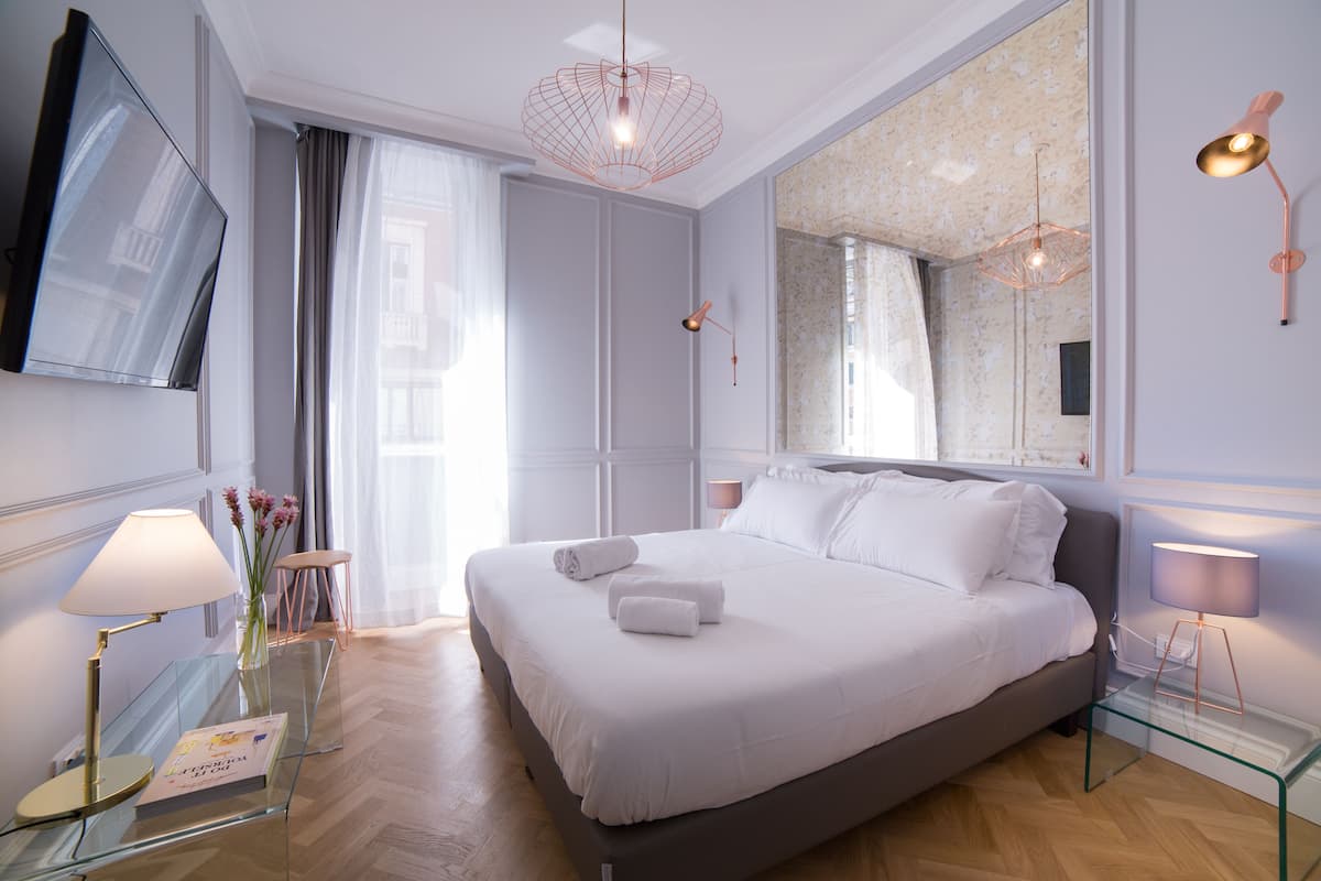 A modern bedroom with a large bed, a flat-screen TV, a glass coffee table, and a mirror.