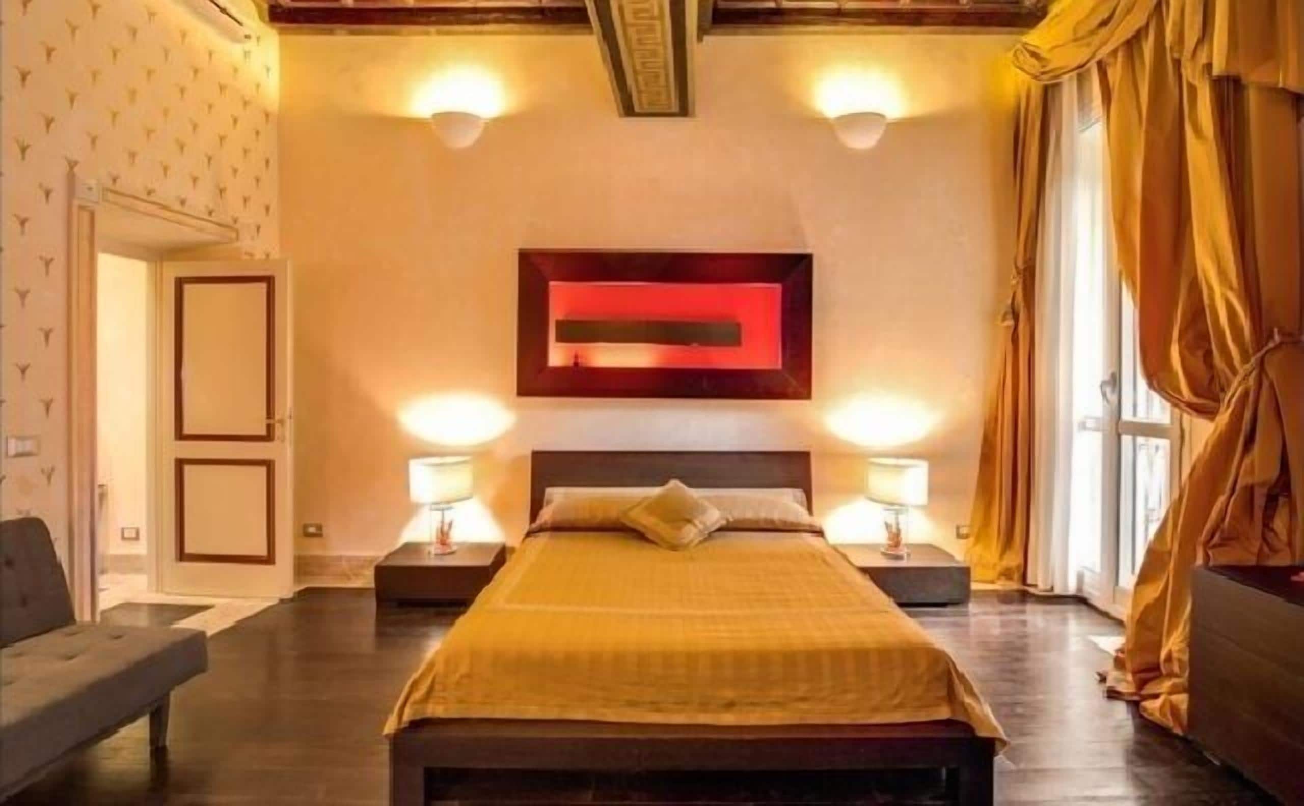 Une chambre à coucher avec un grand lit, deux tables de chevet, une lampe fixée au mur et une fenêtre avec des rideaux.