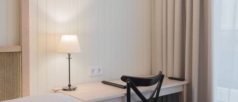 A hotel room with a bed, a desk with a lamp, a chair, and a window with curtains.