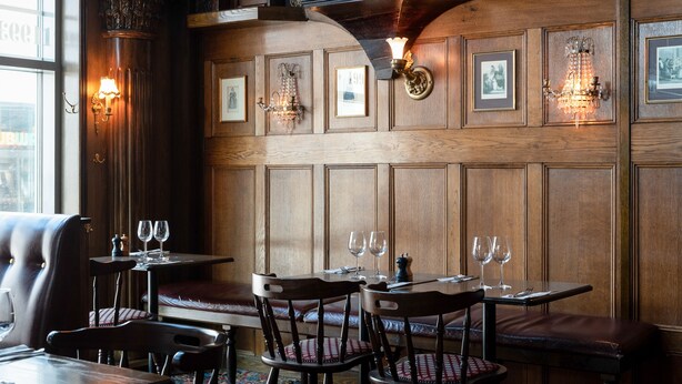 A restaurant with wooden paneling, framed pictures, and candlelit wall sconces. Tables are set with wine glasses and cutlery.