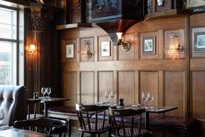 Un restaurant aux murs lambrés, orné de tableaux encadrés et éclairé par des appliques murales à la lumière des bougies. Les tables sont dressées avec des verres à vin et des couverts.
