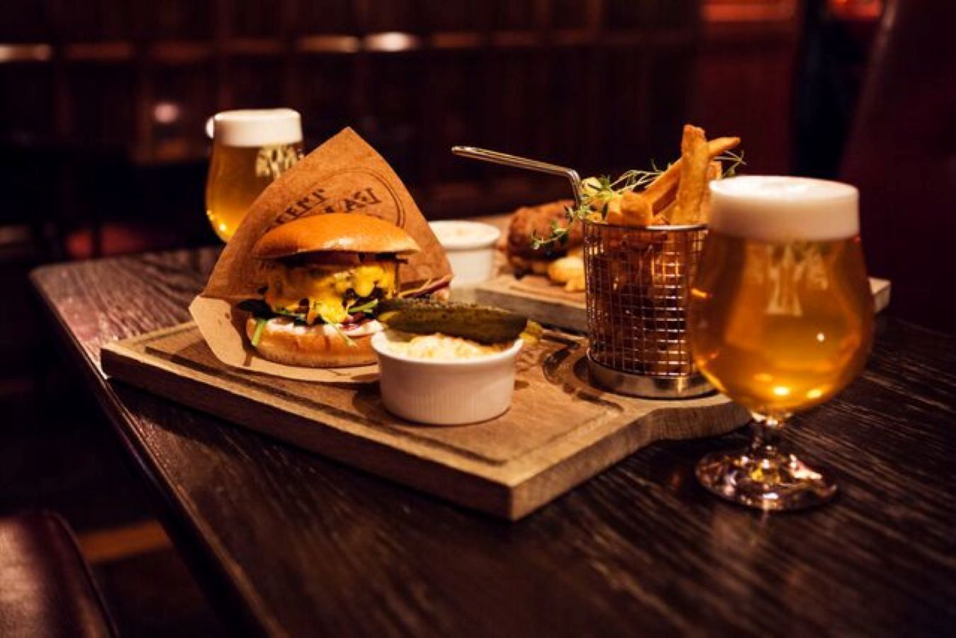 Un vassoio con un hamburger, un sottaceto, una piccola ciotola di insalata di cavolo e un bicchiere di birra su un tavolo di legno.