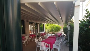 Un patio al aire libre con mesas y sillas, cubierto por un toldo blanco, rodeado de vegetación.