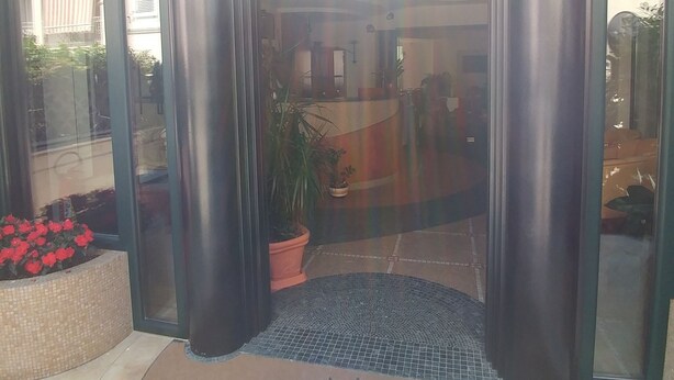 Entrada de un hotel con felpudo que dice "Hotel La Perla", puertas de vidrio grandes y suelo de baldosas.