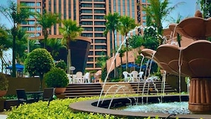 Um hotel moderno com um edifício alto e vários janelões. À frente, encontra-se um jardim ajardinado com uma fonte e vegetação bem cuidada.