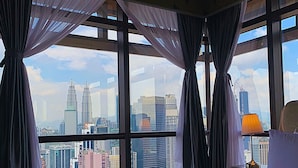 Et hotelværelse med en stor seng, et natbord med lampe, og udsigt til bybilledet inklusive Petronas Towers.