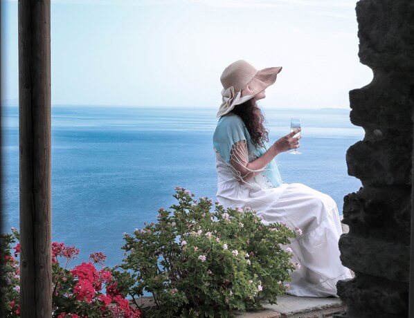 A person sitting by a window overlooking a body of water, wearing a hat and holding a glass. - Aegean Castle  Andros - Adults Only (Andros)