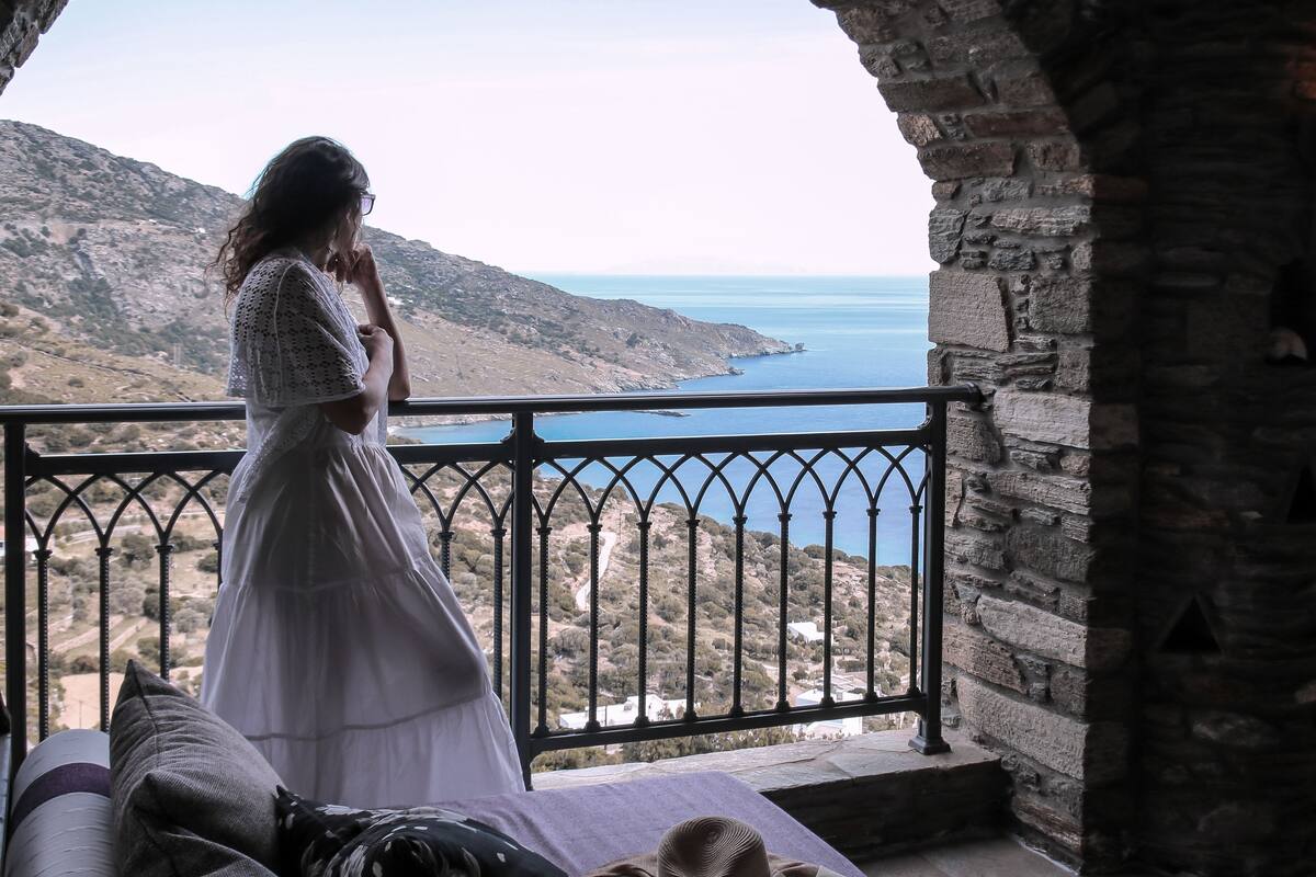 A person in a white dress standing on a balcony with a view of the sea and hills.