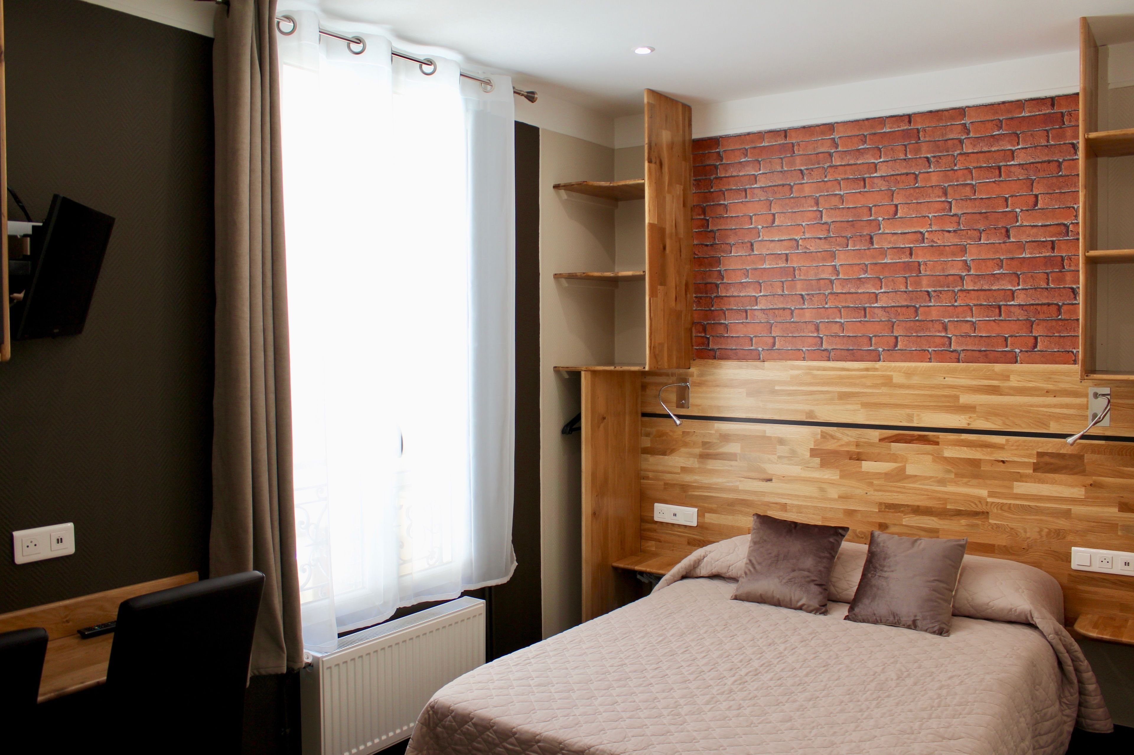 A bedroom with a brick-patterned wall, a bed with a beige quilt, a wooden shelf unit, and a window with curtains.