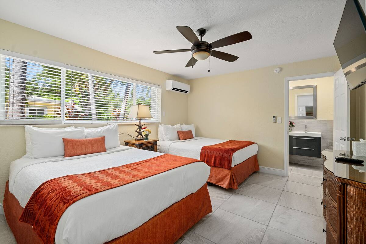 A hotel room with two beds, a ceiling fan, a window with blinds, a bathroom with a sink and mirror, and a view of the outdoors.