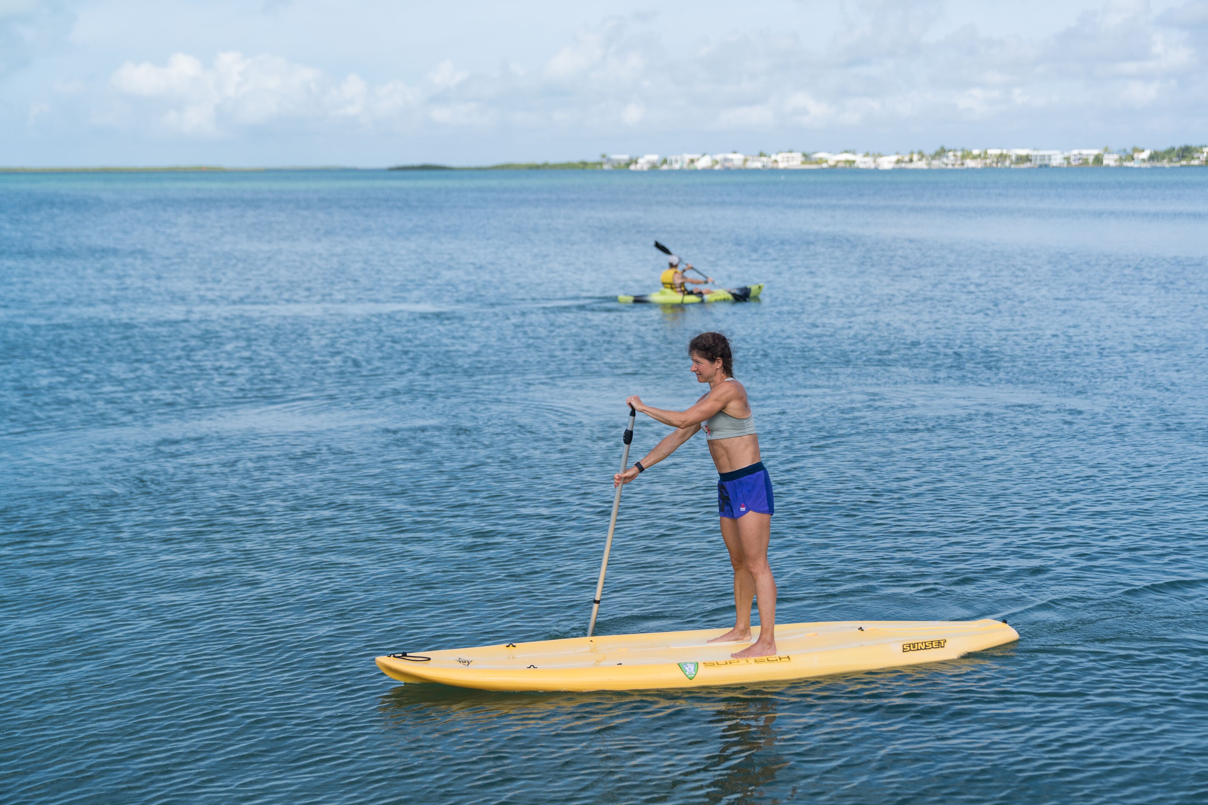 A person paddleboarding on a calm body of water with a distant kayaker.