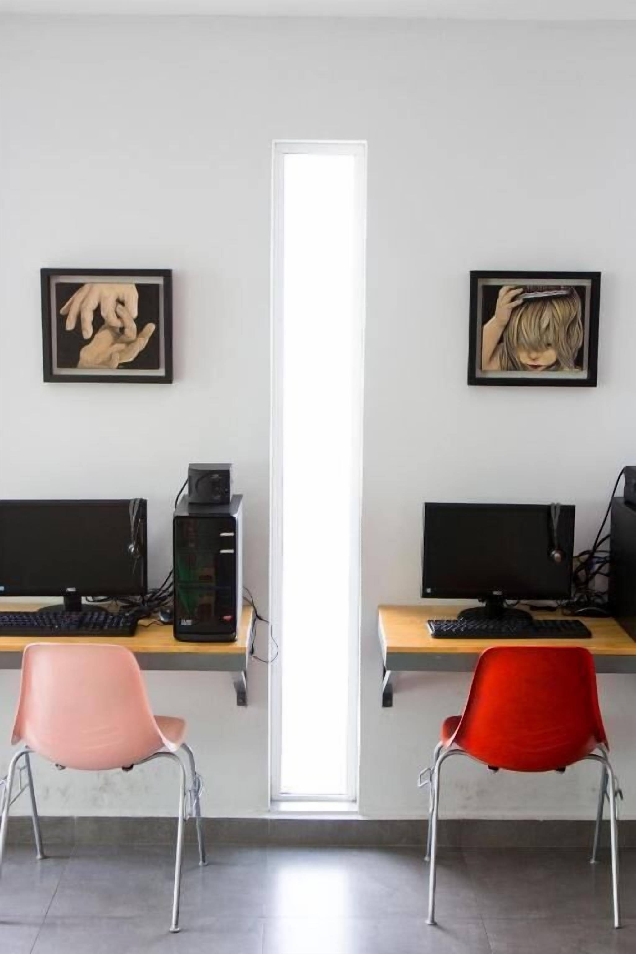 A modern office space with two desks, each with a computer monitor, keyboard, and a small printer. There are two chairs, one pink and one red, in front of the desks. Two framed artworks are hung on the wall above the desks.
