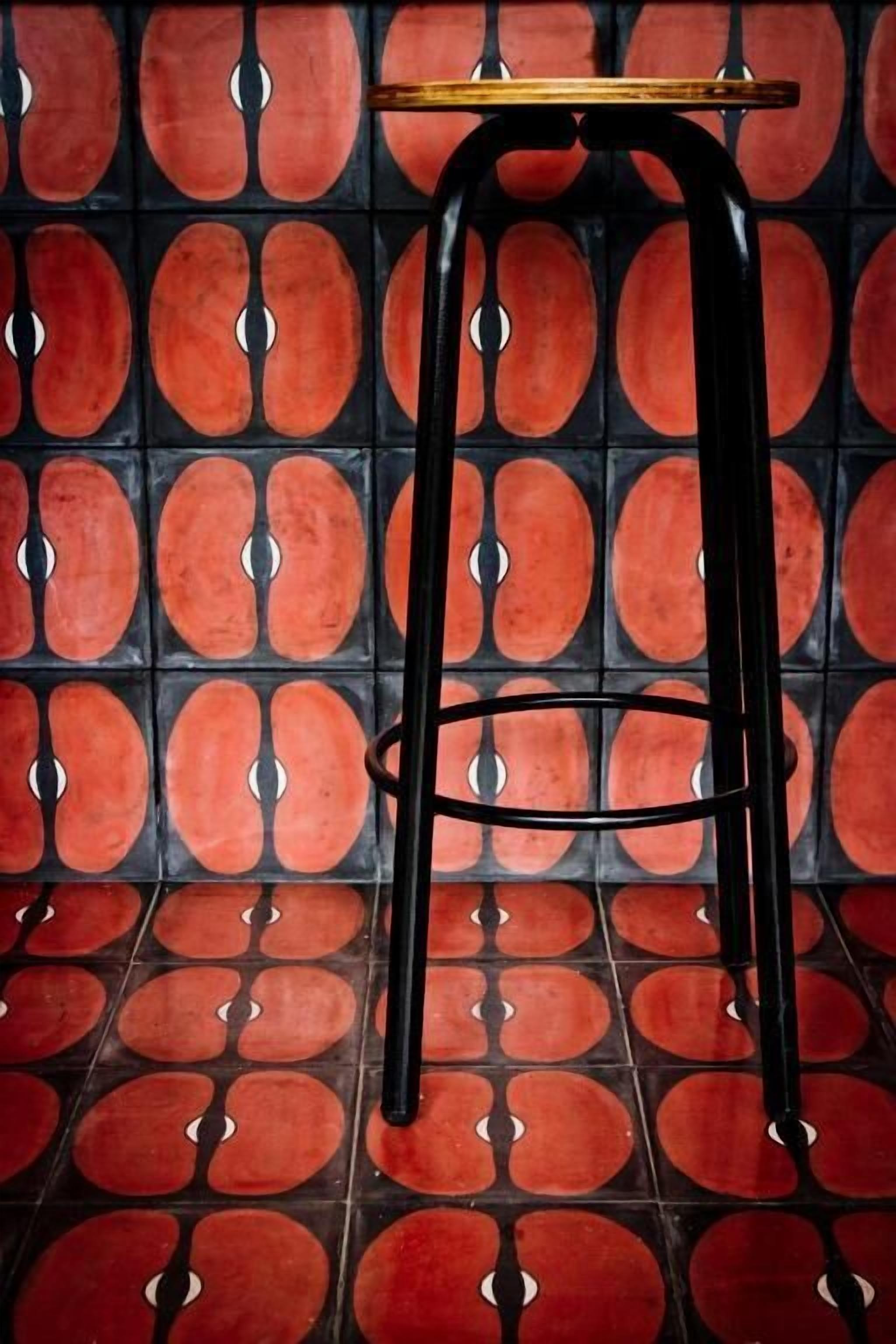 A black metal stool with a wooden top, placed on a tiled floor with a repeating pattern of red and black tiles.