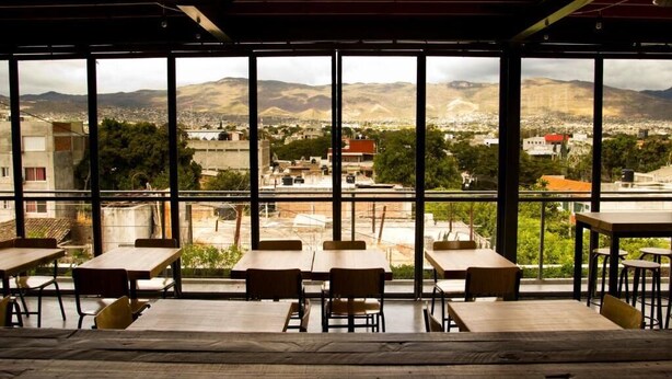 A restaurant with large windows offering a view of a city and mountains. Tables and chairs are arranged for dining. - Hostal Tres Central (Tuxtla Gutierrez)