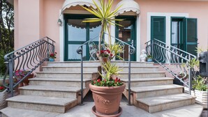 Eine Reihe aus Steinstufen führt zu einem Hauseingang mit grünen Fensterläden, einem Blumentopf mit roten Blüten und einem Metallgeländer.