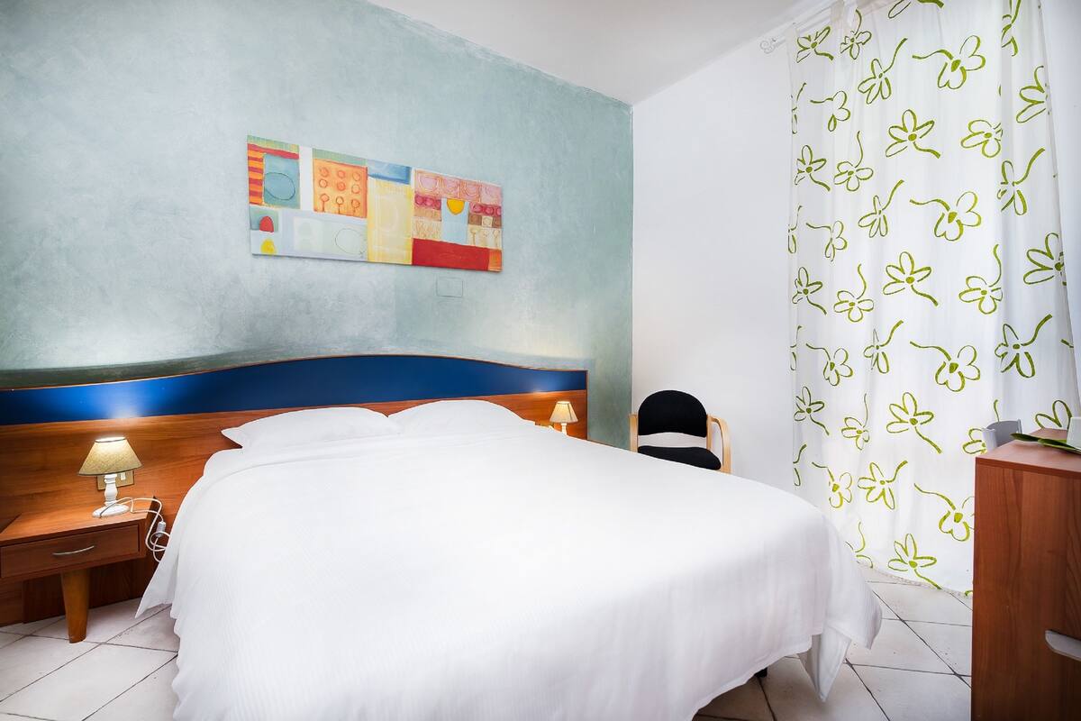 A hotel room with a bed, a nightstand with a lamp, a chair, and a wall with a floral curtain.