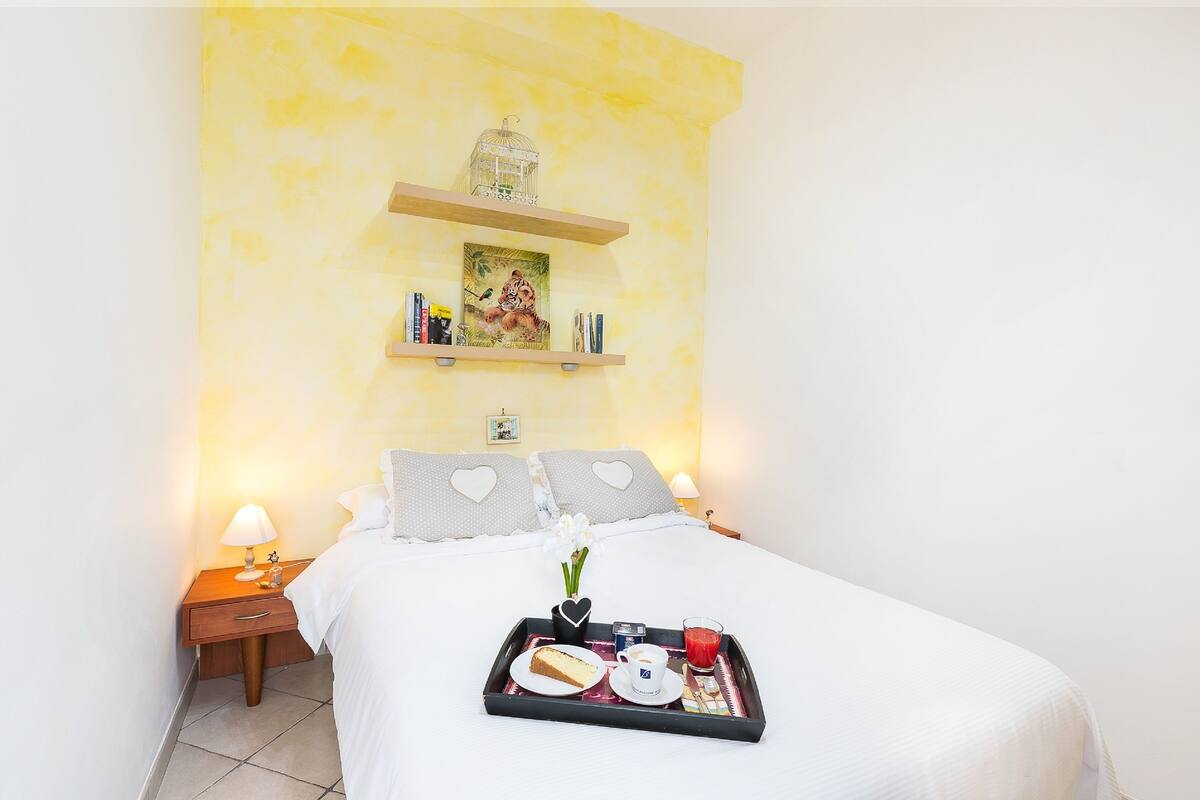 A neatly made bed with a tray of breakfast items, a nightstand with a lamp, and a shelf with decorative items.