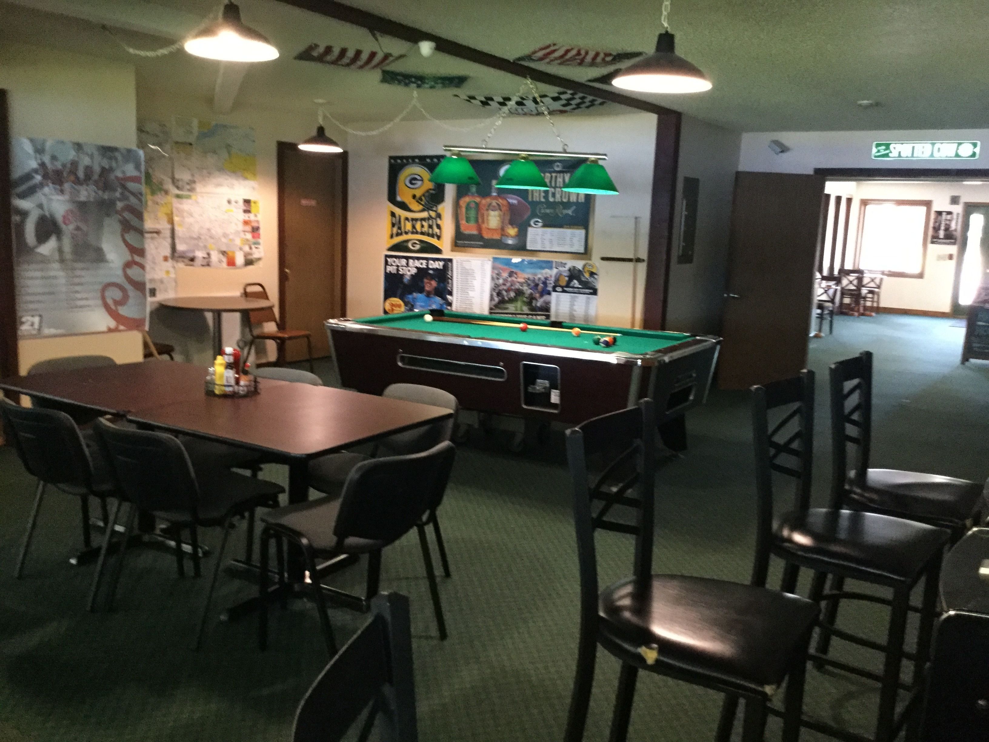 Une table de billard dans une pièce entourée de chaises, une table avec des bouteilles et des affiches accrochées au mur.
