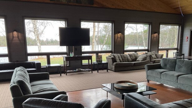 A spacious living room with leather sofas, a coffee table, and a flat-screen TV. Large windows offer a view of the outdoors.