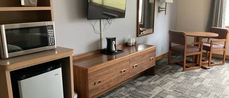 A hotel room with a wooden desk, a microwave, a television, a small refrigerator, a coffee maker, a mirror, and a chair.