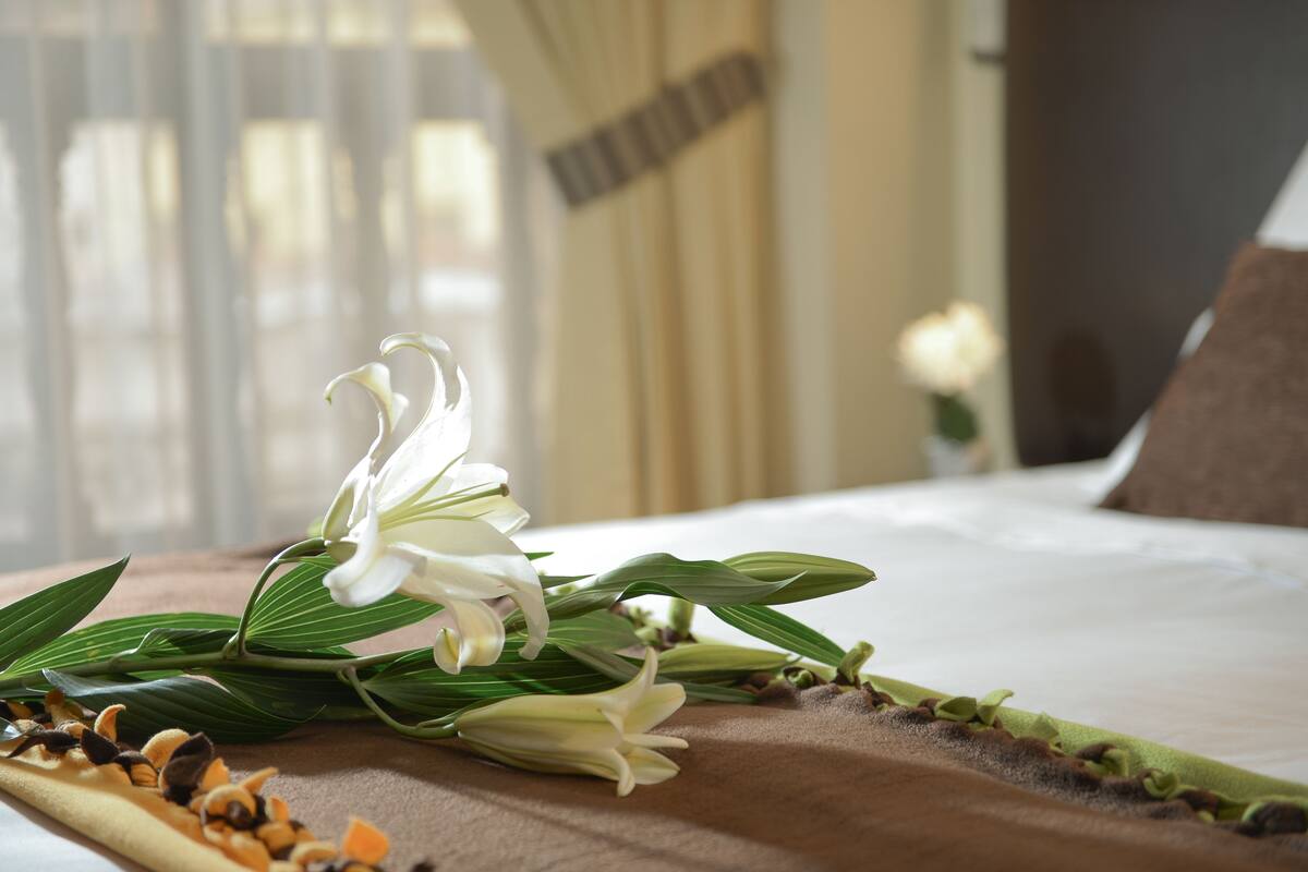 Un lit avec du linge de lit blanc, un coussin décoratif marron et un bouquet de lys blancs posé sur un tissu marron.