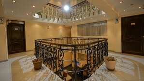 Um hall de hotel com uma escadaria central, uma receção e uma grande janela com cortinas.