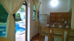 Una habitación con una mesa de comedor preparada para cuatro personas, un refrigerador, una ventana con cortinas y vista a una piscina.