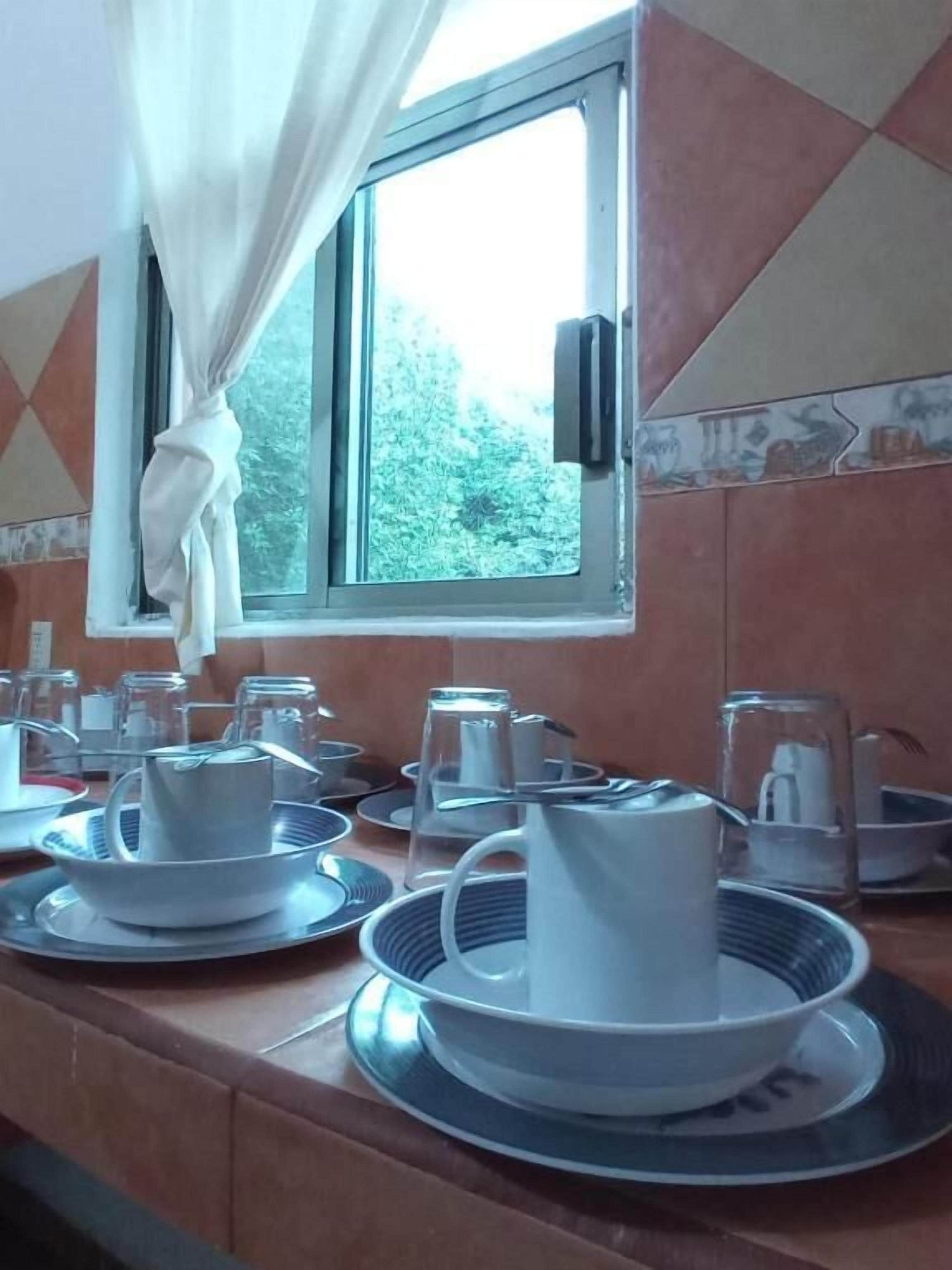 A breakfast setup with plates, cups, and a pitcher on a counter by a window with curtains.