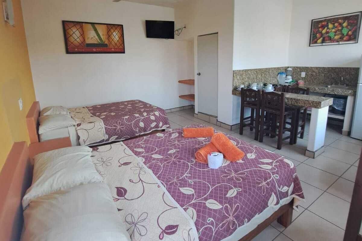 A hotel room with two beds, a kitchenette, a TV, and artwork on the walls.