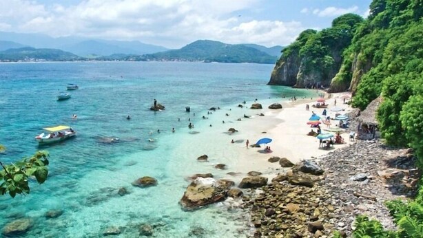 Una playa con aguas turquesas cristalinas, costa rocosa y abundante vegetación.