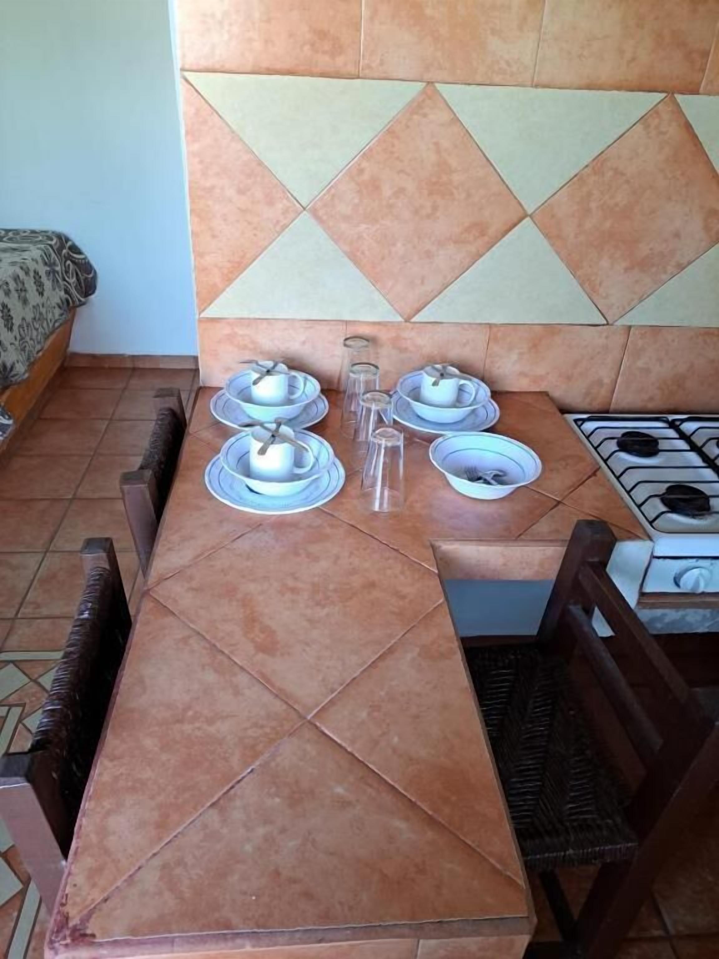 Een eetgedeelte met een betegelde tafel gedekt voor vier personen, een gasfornuis en een gestreept bankje op de achtergrond.