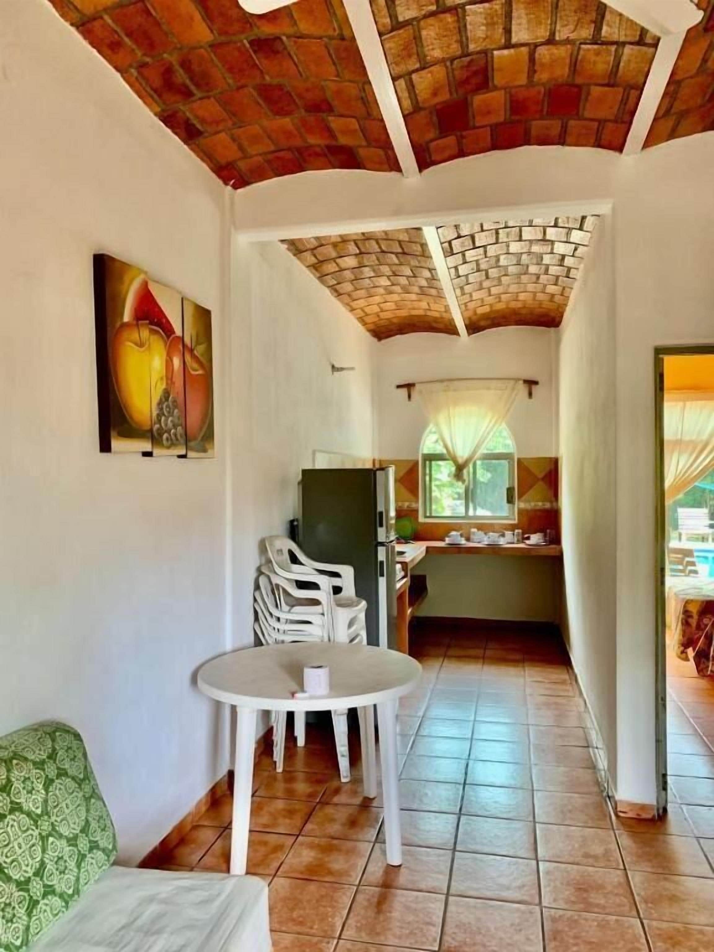 A room with a dining table and chairs, a kitchen area with a window, and a brick ceiling.