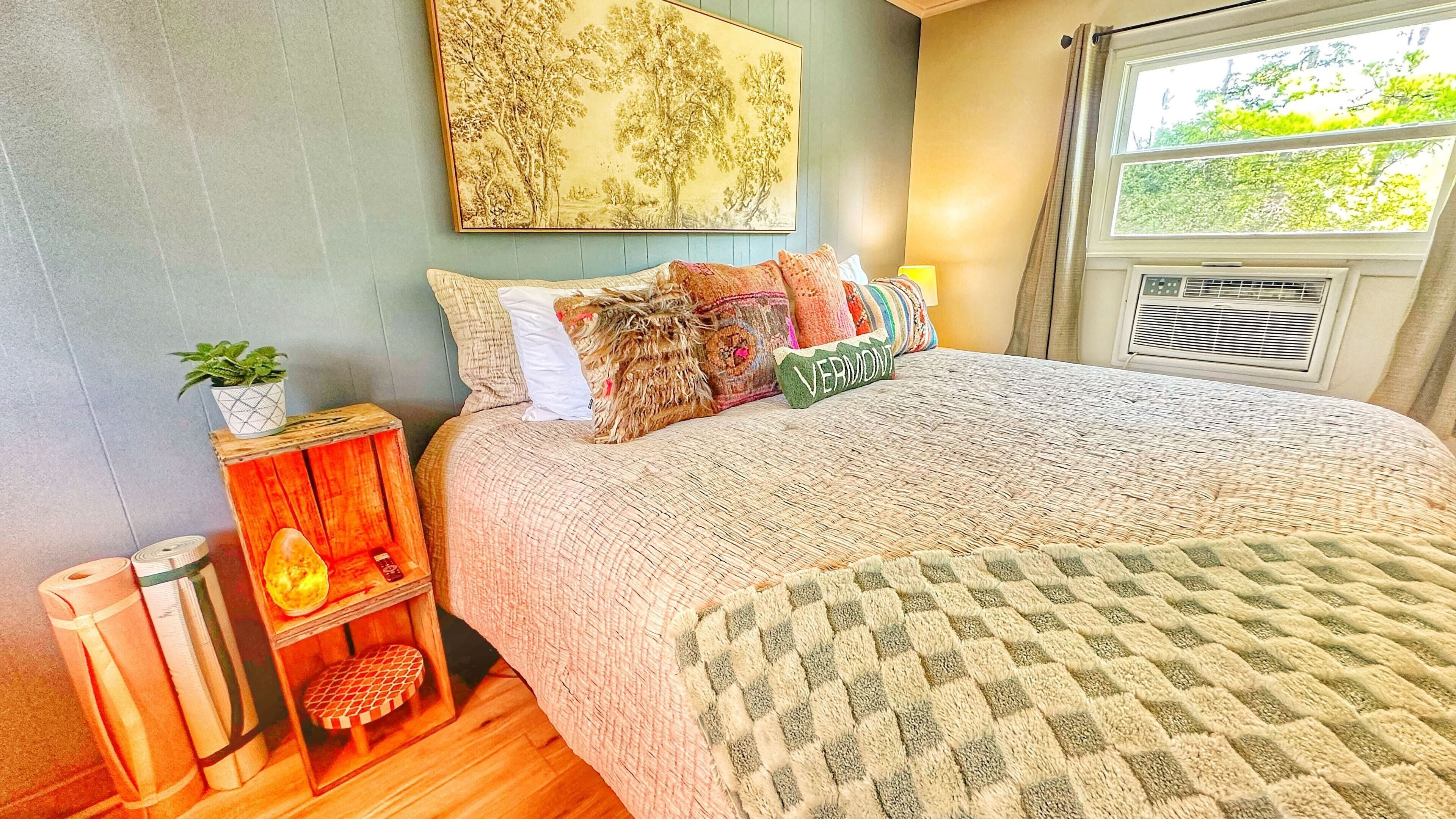 A neatly arranged bedroom with a large bed, a bedside table with a lamp, a small shelf with a plant, and a window with a view of greenery.