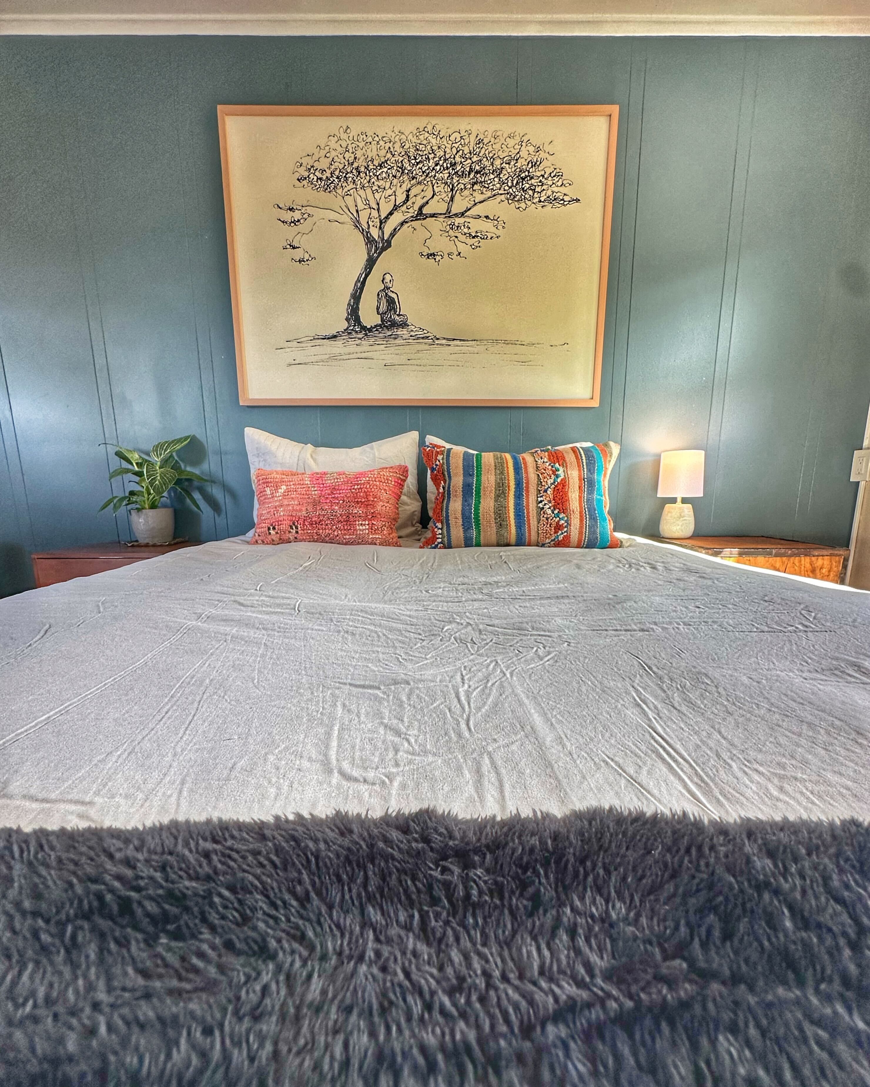 A large bed with a fluffy gray rug, a framed tree painting on the wall, and a bedside lamp.