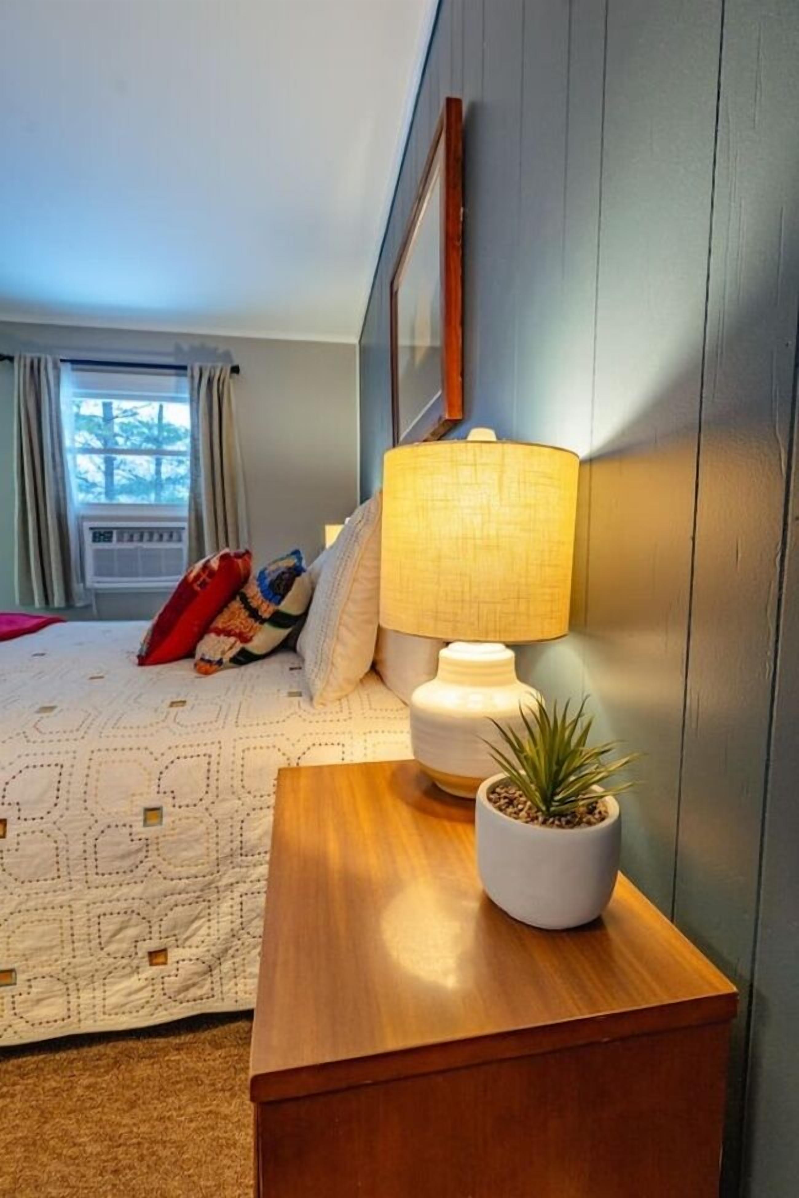 Une chambre à coucher avec un lit, une table de chevet avec une lampe et une plante en pot, une fenêtre avec des rideaux et un tableau encadré au mur.