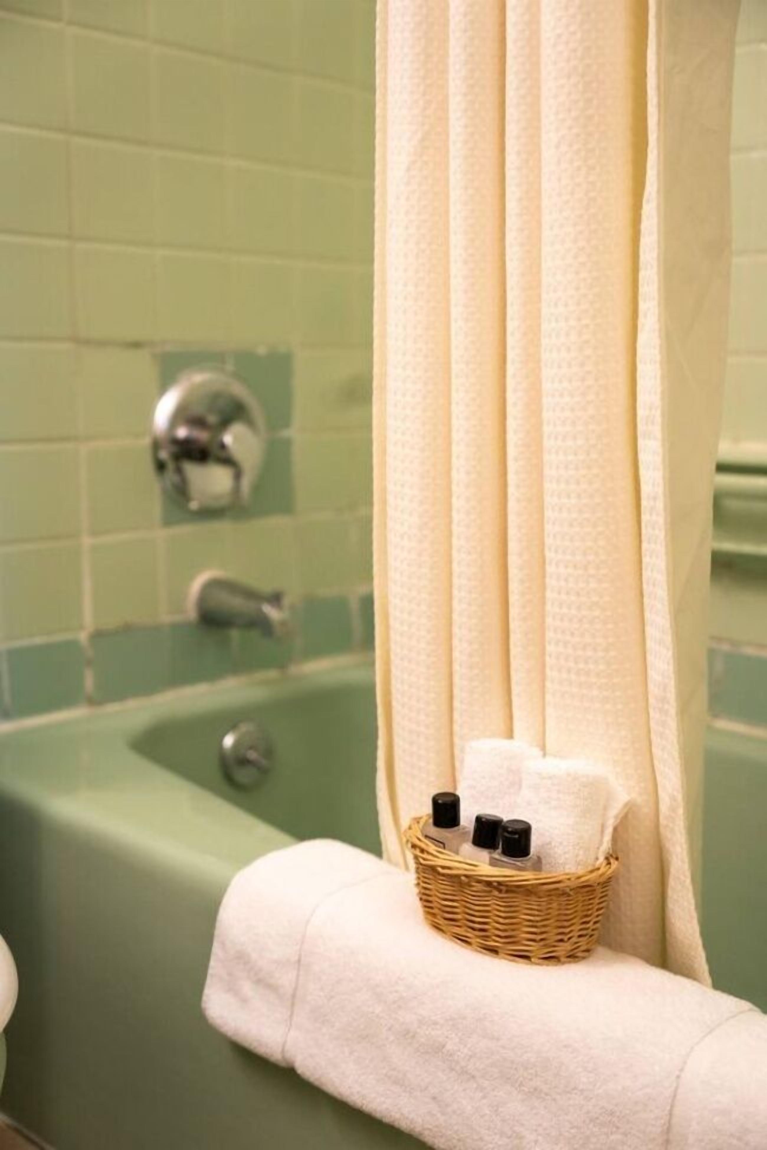 Ein Badezimmer mit grünen Fliesen, einer Badewanne, einem weißen Handtuch und einem Korbwannen-Set mit Toiletteartikeln.