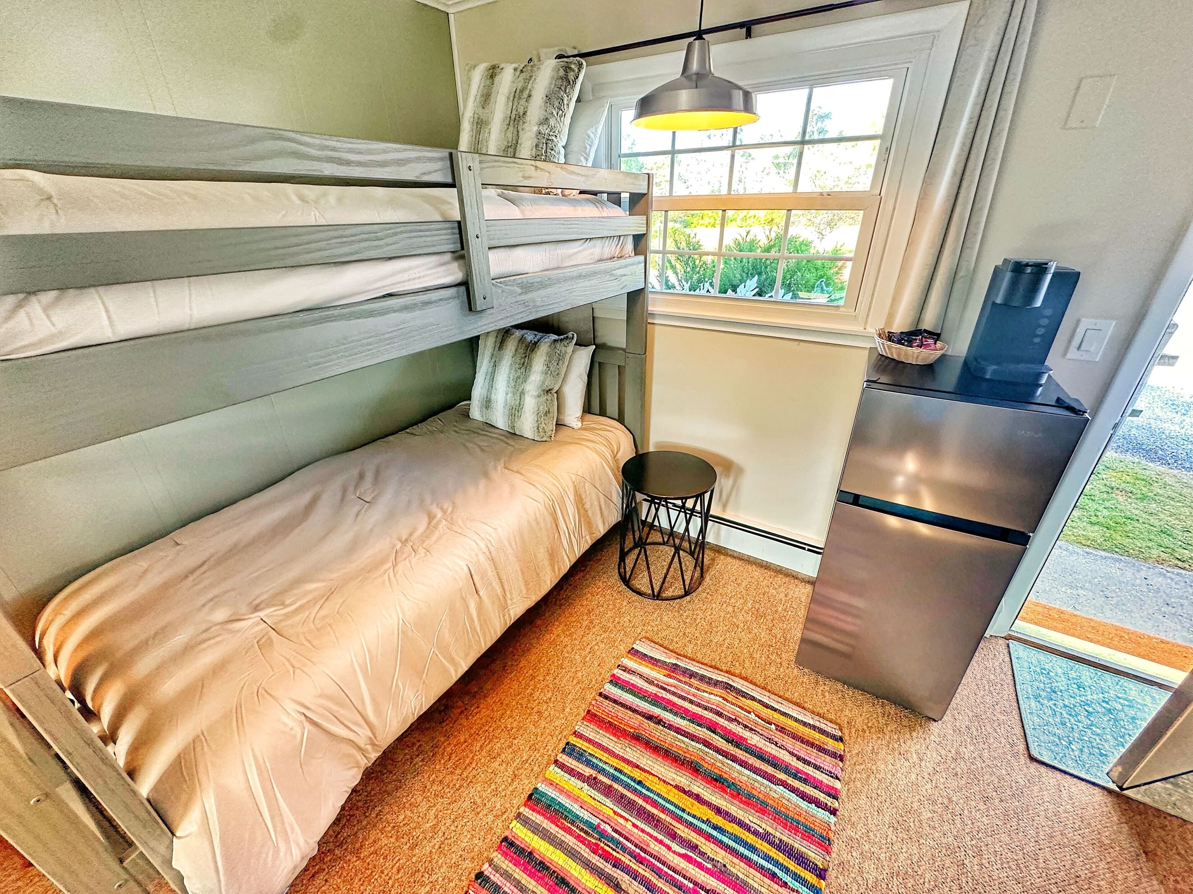 A compact room with bunk beds, a small round table, a colorful striped rug, a refrigerator, and a window with a view of greenery.