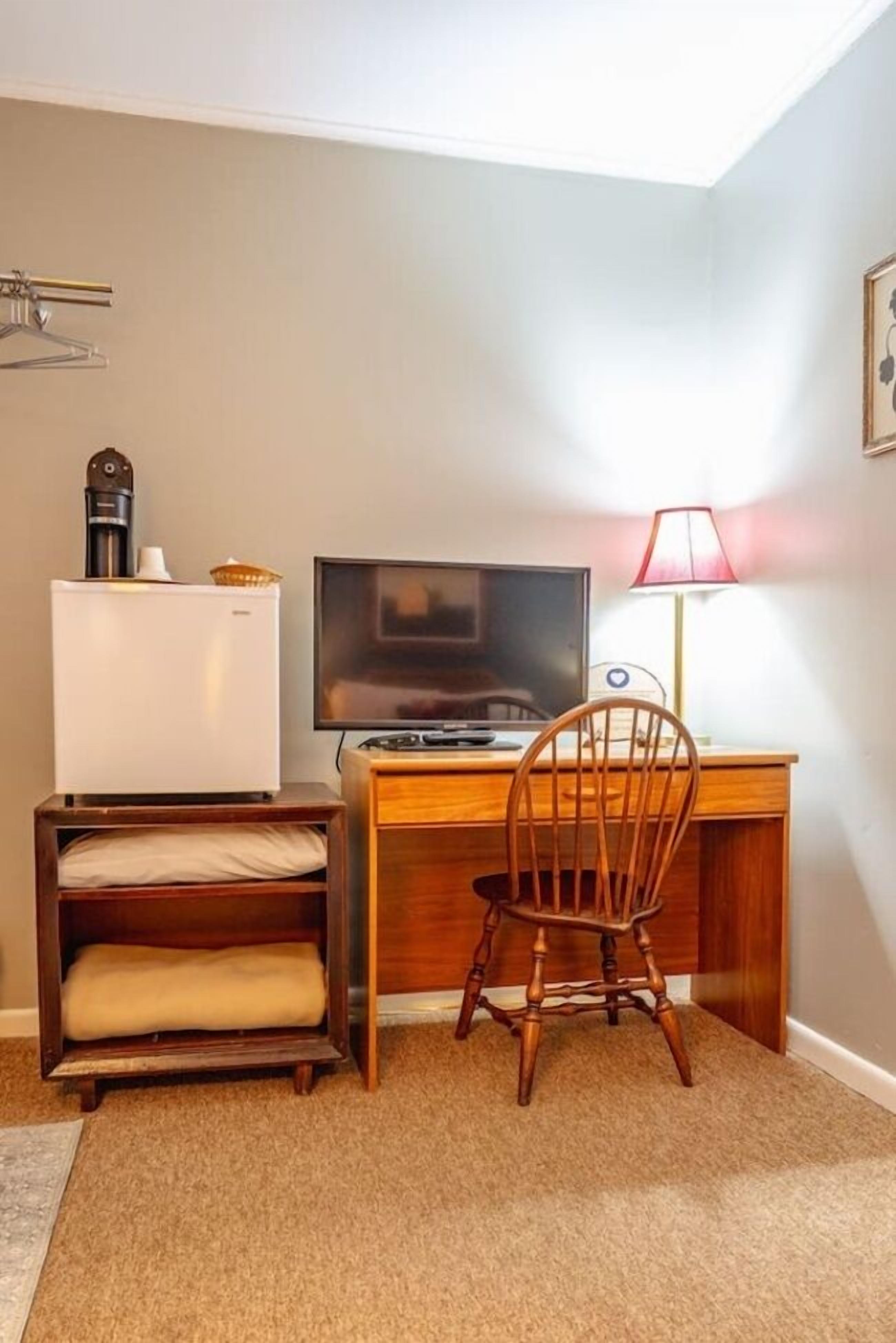 A compact room with a wooden desk, a chair, a television, a refrigerator, and a lamp.